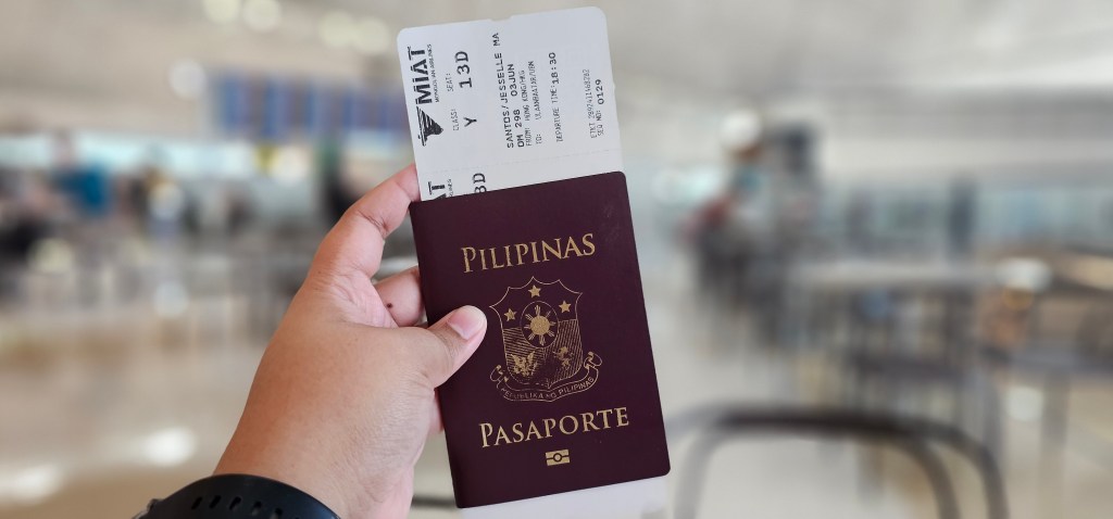 A hand holding a Philippine passport and a boarding pass at an airport, with a blurred terminal background.