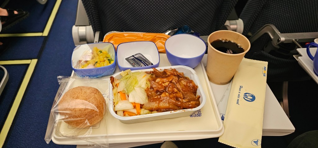 A tray of airplane meal featuring chicken in sauce with vegetables, a side of whole grain roll, a fruit salad, a small cup, and a paper cup of coffee, set on an airplane seat.