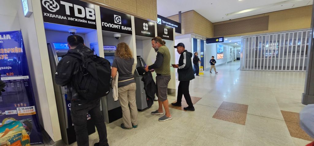 People using ATMs at an airport terminal, with multiple bank machines visible, and travelers in the background.