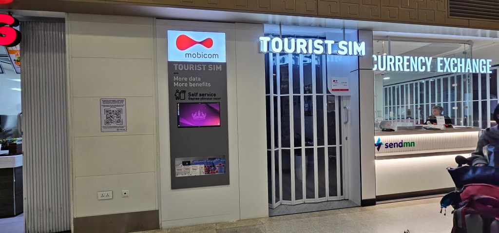 Exterior view of a Mobicom sign featuring 'TOURIST SIM' with information about self-service and benefits, next to a currency exchange office.