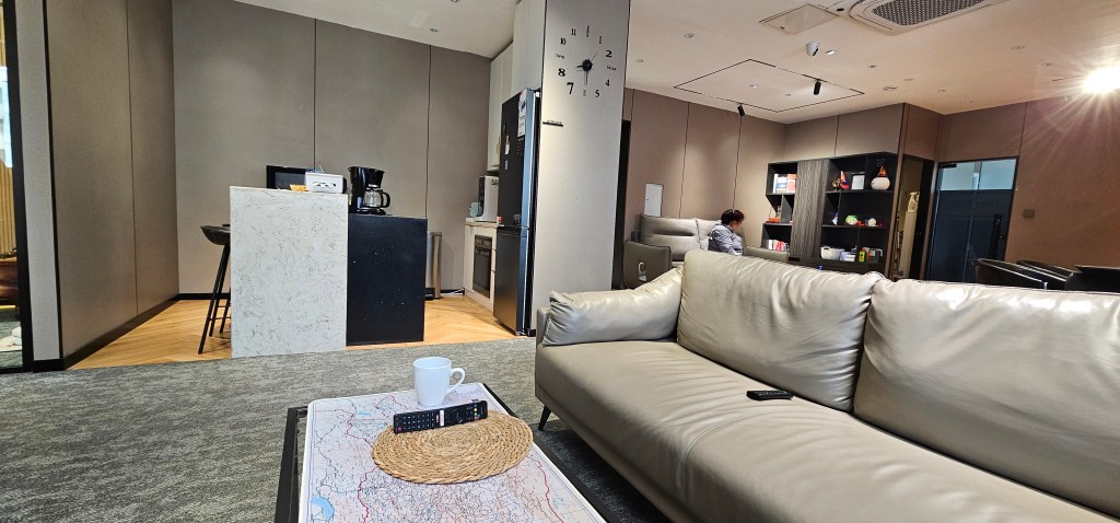 A modern living room with a light-colored leather sofa, a coffee table featuring a map and remote control, and a kitchen area in the background. A person is seen working at the kitchen counter.