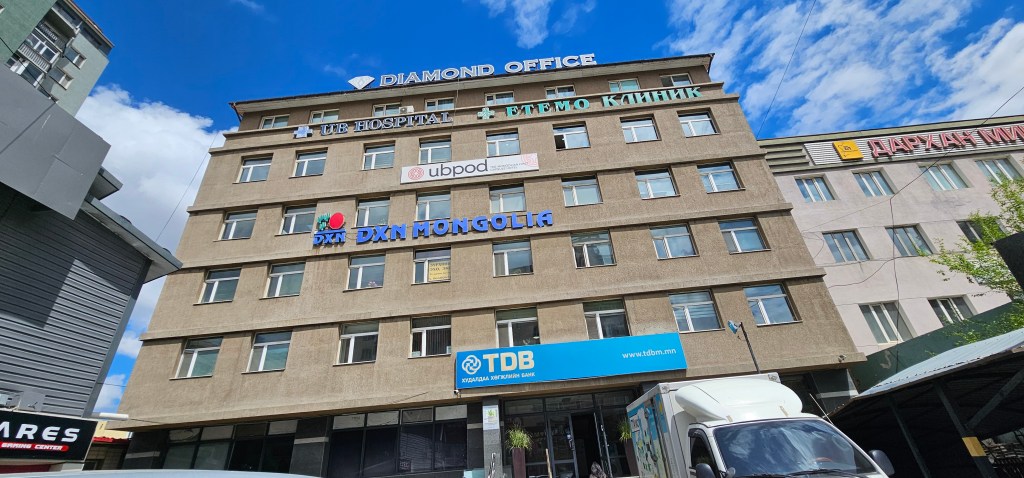 Exterior view of a multi-story building with various signs including 'Diamond Office', 'UB Hospital', and 'DXN Mongolia'. The sky is partly cloudy.