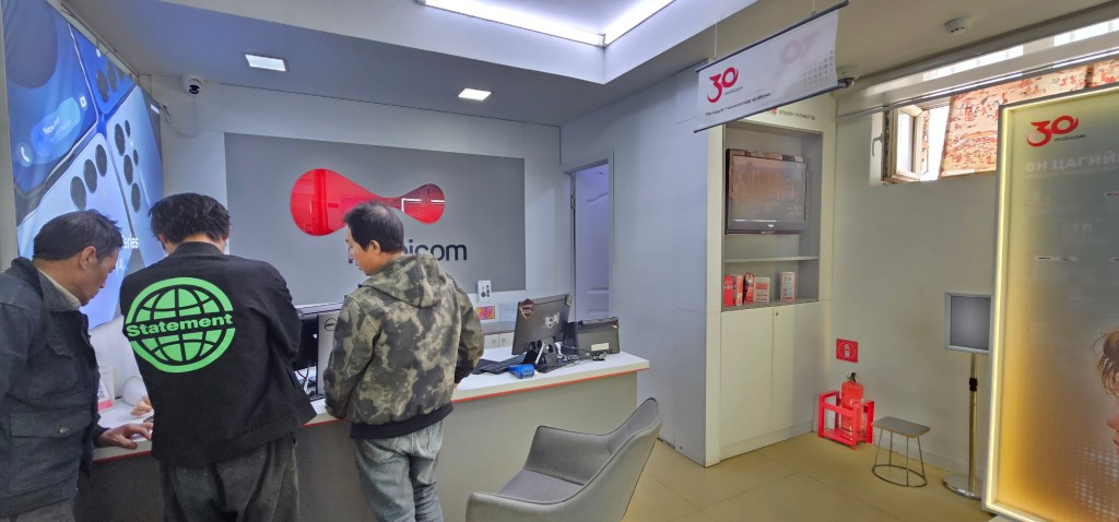 Interior of a mobile service store with three customers interacting at the counter and various display items in the background.