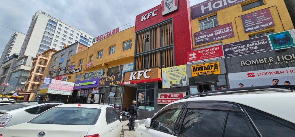 View of a street in an urban area featuring commercial buildings, including a prominent KFC restaurant, various shop signs, and parked cars.