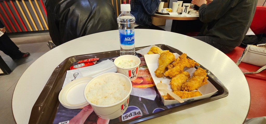 A tray filled with crispy fried chicken pieces, a serving of rice, a side of coleslaw, and a bottle of water, set on a table in a casual dining environment.