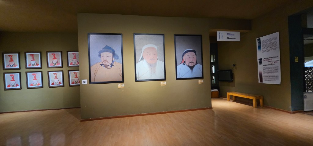 An indoor exhibit featuring framed portraits of historical figures on a green wall, with additional framed artworks on the left and an informational display on the right.