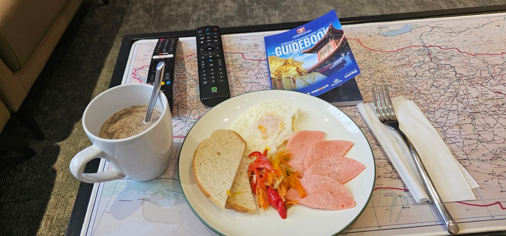 A plate of breakfast consisting of fried eggs, slices of processed meat, and sautéed vegetables, accompanied by a slice of bread. A cup of coffee is next to the plate, with a remote control, a fork, and a guidebook placed on a map-covered table.