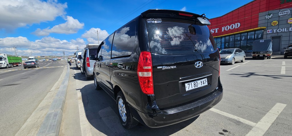 View of a black Hyundai Starex parked along a roadside with other vehicles in the background and a fast food restaurant nearby.