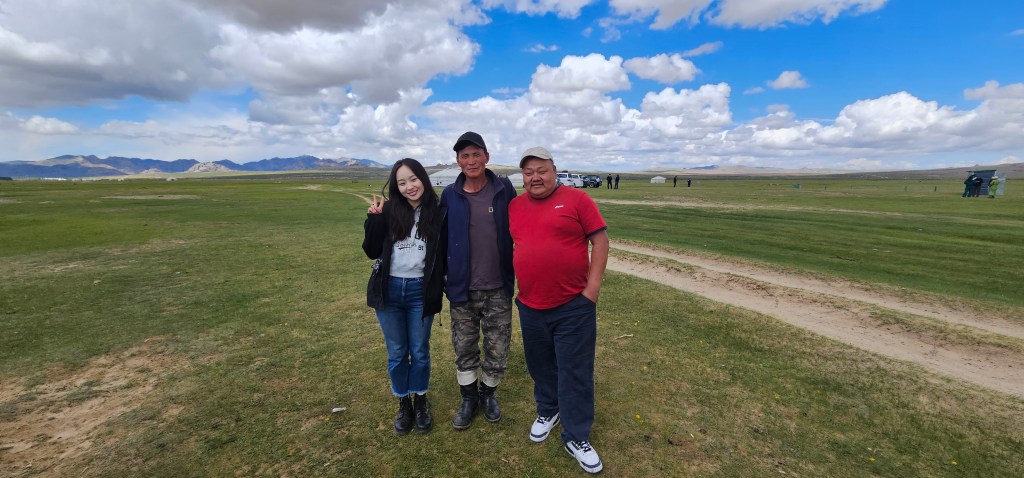 Three individuals pose together outdoors on a grassy landscape under a blue sky with clouds.