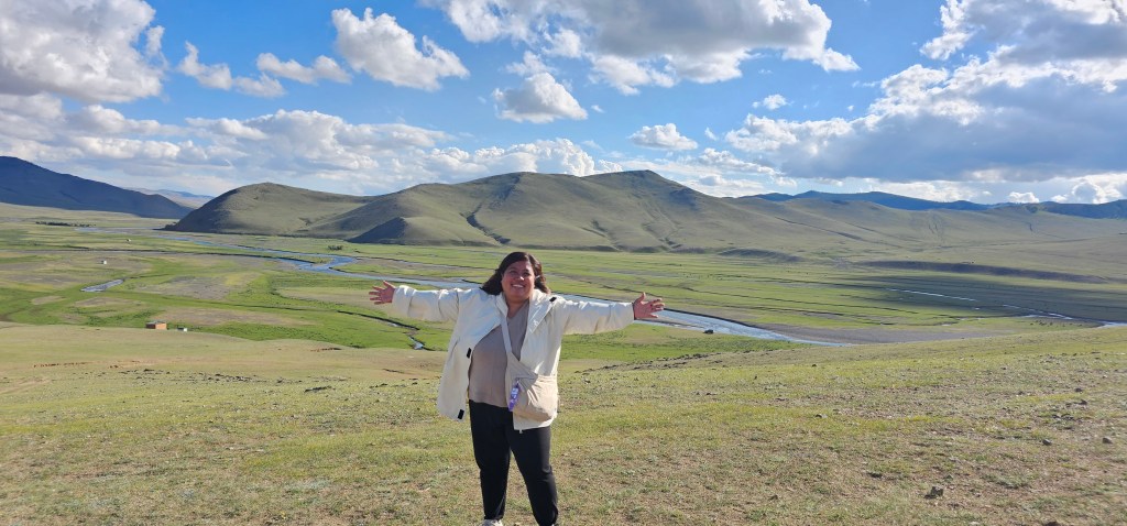 A cheerful woman standing on a grassy hillside with arms outstretched, overlooking a scenic landscape of rolling hills and a winding river under a blue sky with fluffy white clouds.