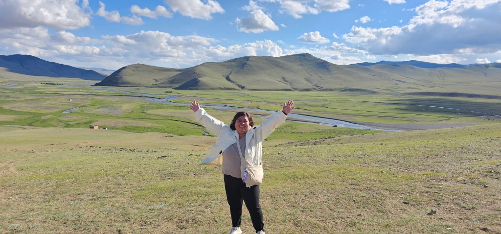 A person stands joyfully in a vast green landscape with rolling hills and a river flowing in the background under a blue sky with scattered clouds.