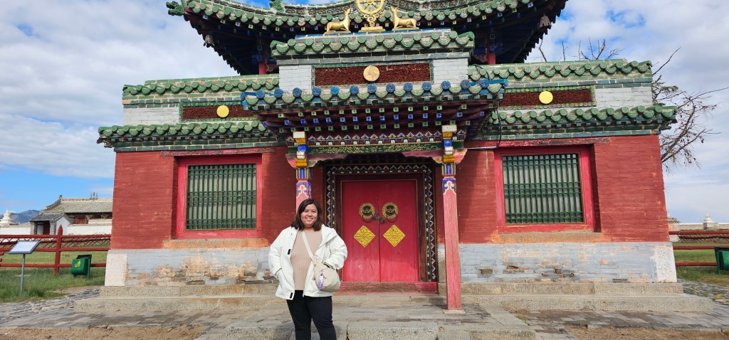 A woman stands in front of a colorful traditional building with ornate architecture, featuring a red facade and green roof decorations, set against a partly cloudy sky.