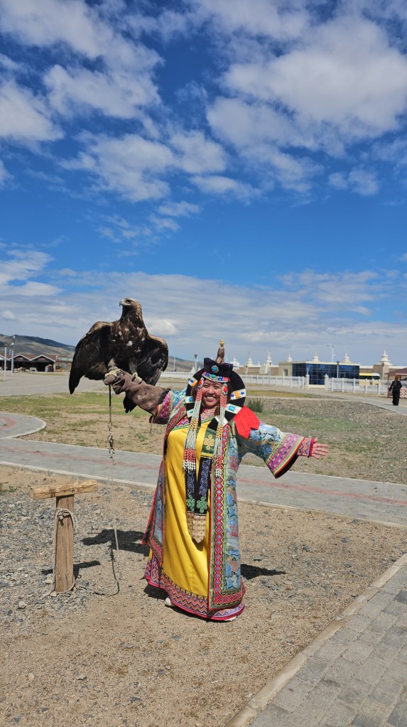 A person dressed in vibrant traditional attire stands outdoors with an eagle perched on their arm, smiling against a backdrop of blue sky and fluffy clouds.