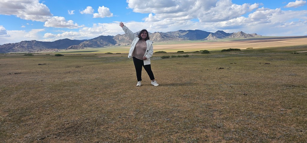 A woman stands in a vast, open landscape with mountains in the background, smiling and gesturing cheerfully. The scenery features grassy terrain under a bright blue sky filled with clouds.