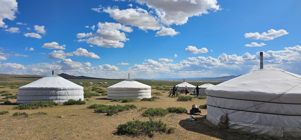 A picturesque landscape featuring several white yurts against a blue sky with fluffy clouds. The setting is expansive, showcasing grassy land and distant hills.