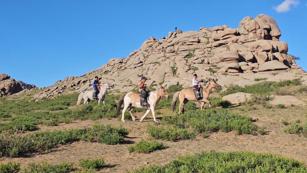 Four people riding horses on a grassy hillside with large rock formations in the background and a clear blue sky.