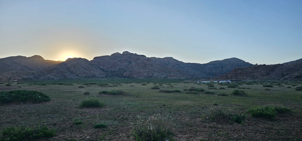 A panoramic view of a mountainous landscape at sunset, featuring grassy plains and sparse vegetation in the foreground, with white tents visible in the distance.
