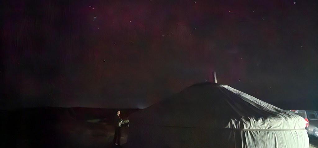 A person stands next to a yurt under a starry night sky, with a car parked nearby.