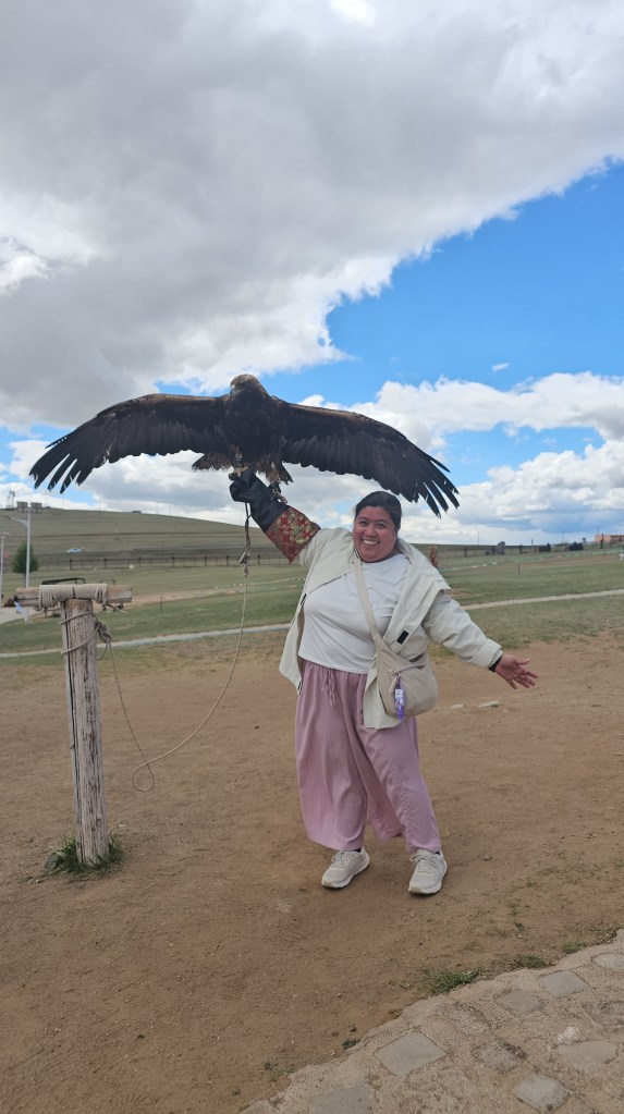 A woman smiling and posing with her arms outstretched, holding a golden eagle on her arm. The background features a grassy area under a partly cloudy sky.