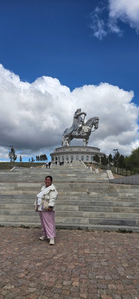 A person stands on stone steps in front of a large, silver equestrian statue against a backdrop of blue sky and fluffy clouds.