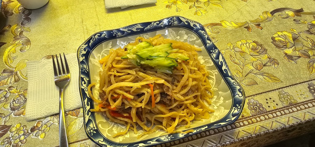 A plate of stir-fried noodles garnished with green cucumber slices, placed on a decorative tablecloth with floral patterns.