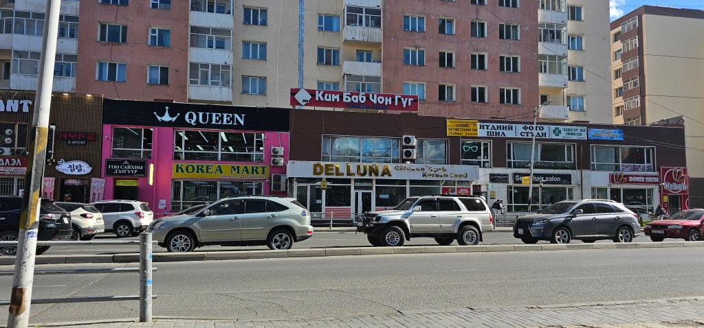 Street view featuring various shops, including a store named 'Korea Mart' and 'DelluNa', with several parked cars and buildings in the background.