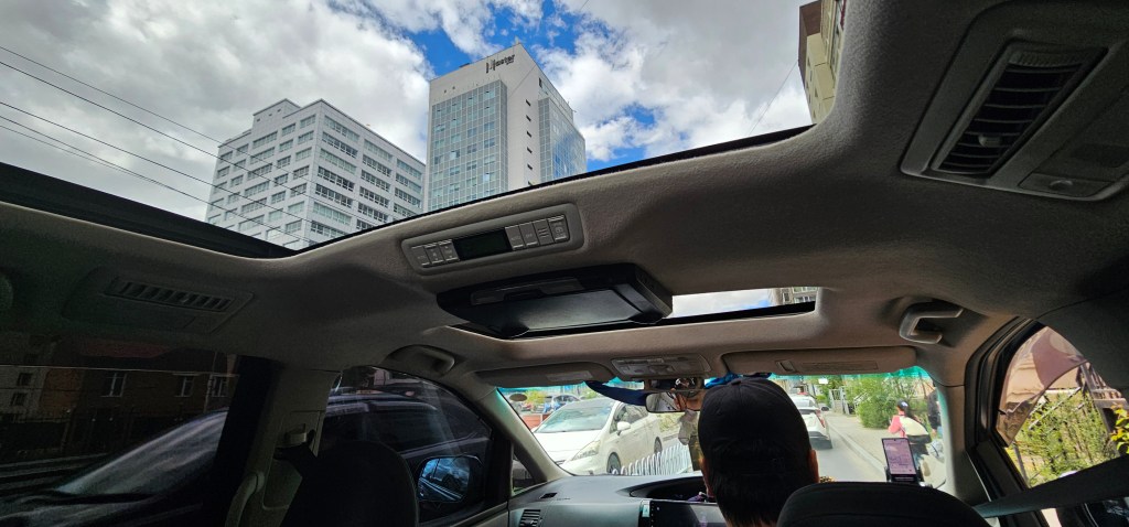 View from inside a car showing a sunroof, the driver's head, and a cityscape with tall buildings and cloudy skies.