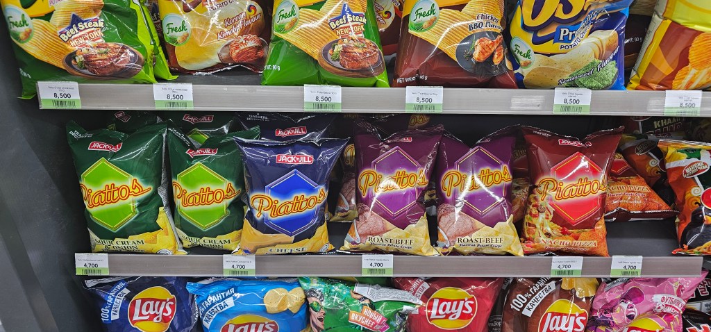 A display of various snack chips in a grocery store, featuring Piattos in flavors like Sour Cream and Cheese, Roast Beef, and Nacho Pizza, along with other brands like Lay's and Oishi.