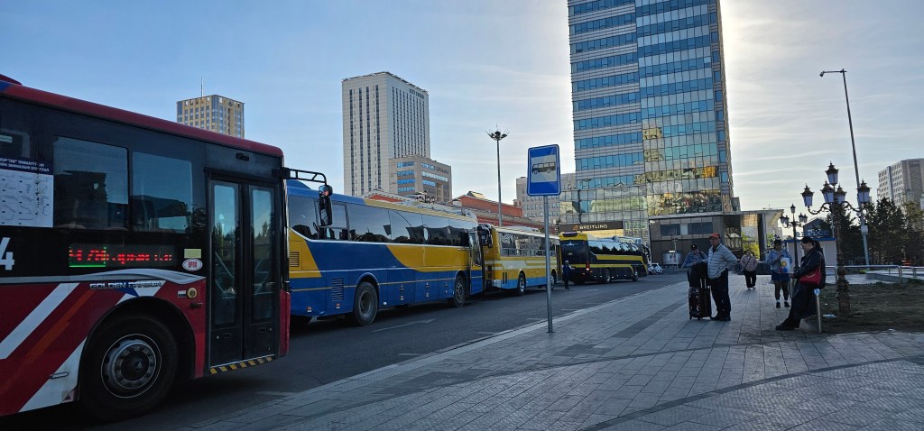 A busy urban bus stop featuring several buses lined up, with modern buildings in the background and passengers waiting on the sidewalk.