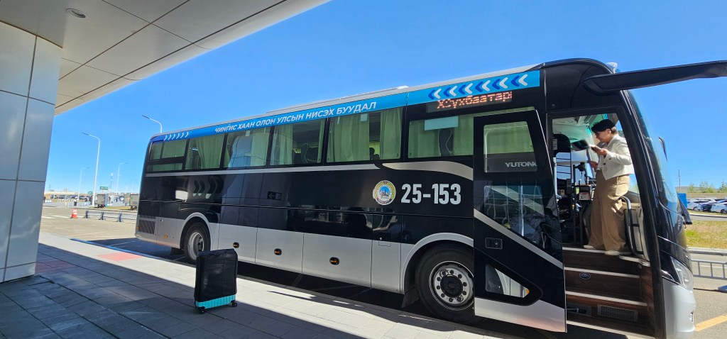 A black and silver public bus parked at a terminal with a person boarding and a suitcase nearby under clear blue skies.