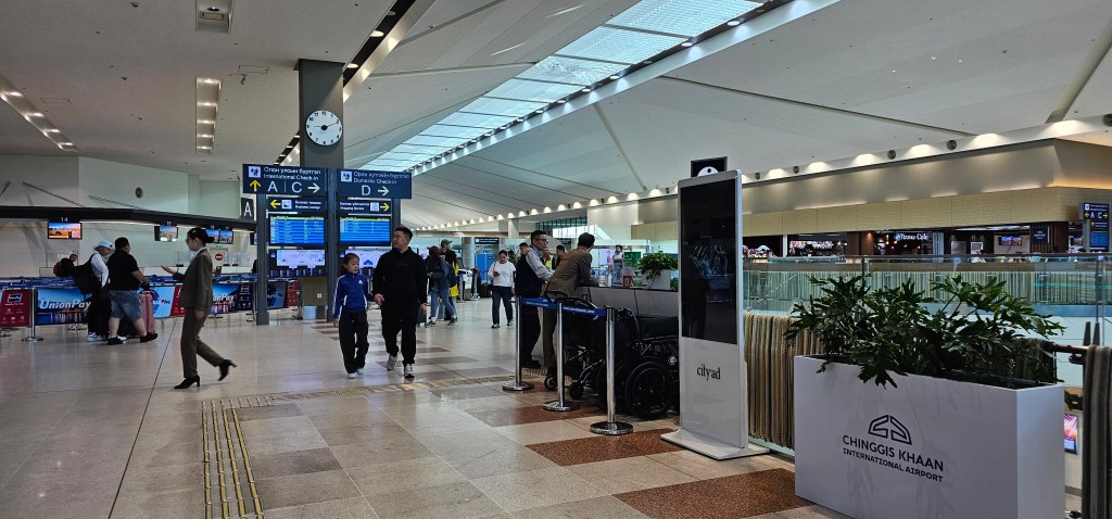Interior view of Chinggis Khaan International Airport with travelers, check-in counters, flight information screens, and plants.