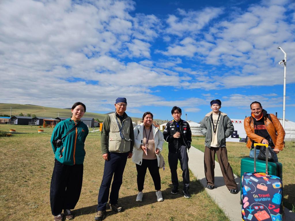 A group of six people stands together outdoors, smiling. They are dressed in casual clothing, with a few carrying bags. The background features a grassy area and gers or yurts, under a partly cloudy blue sky.