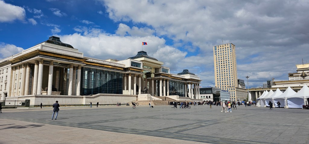 Exterior view of a large historic building with columns and a modern glass structure, set against a blue sky with clouds. People are walking in the surrounding plaza.