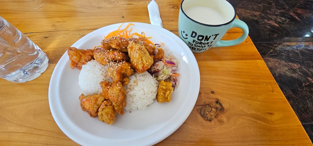 A plate of fried chicken pieces with sesame seeds served over two portions of white rice, accompanied by a small salad and a cup of milk on a wooden table.