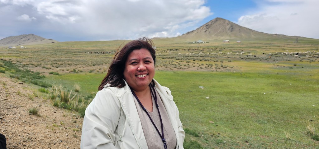 A woman smiling outdoors in a grassy landscape with a mountain in the background under a cloudy sky.