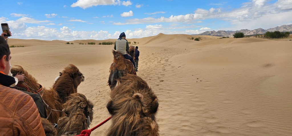 A group of people riding camels through a sandy desert landscape with dunes under a partly cloudy sky.