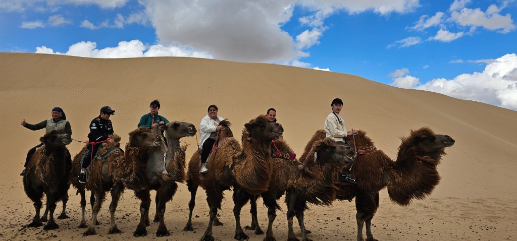 A group of six people riding camels in a desert landscape with sand dunes and a blue sky with clouds.