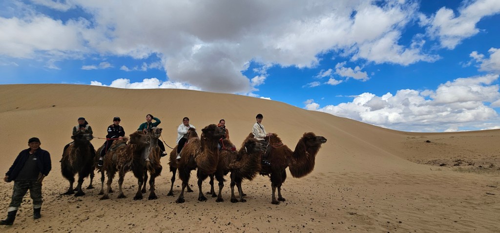 A group of people riding camels in a desert landscape under a blue sky with clouds.