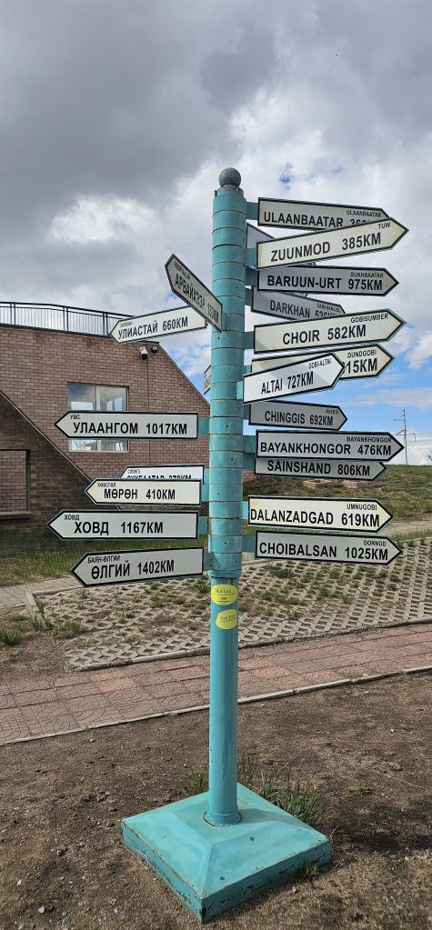 A signpost with multiple directional arrows, indicating distances to various cities in Mongolia, including Ulaanbaatar, Zuunmod, and Darkhan, set against a cloudy sky.