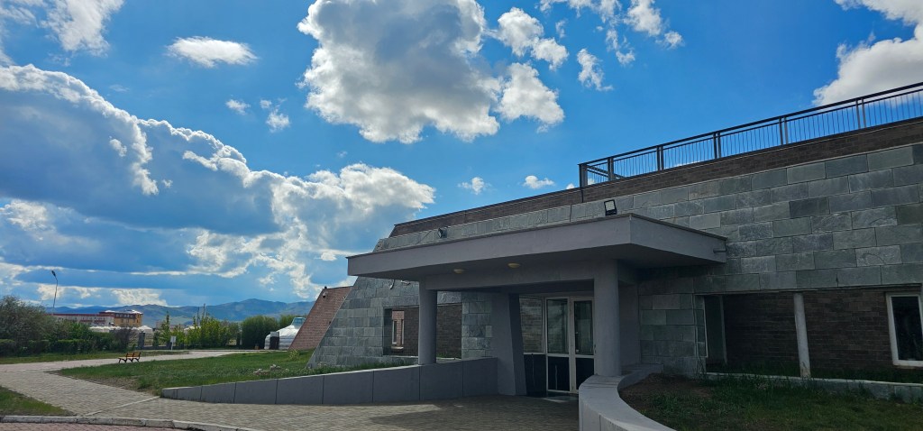 A modern building with a stone exterior, featuring a curved pathway leading to the entrance. The sky is partly cloudy with blue tones and distant mountains visible in the background.