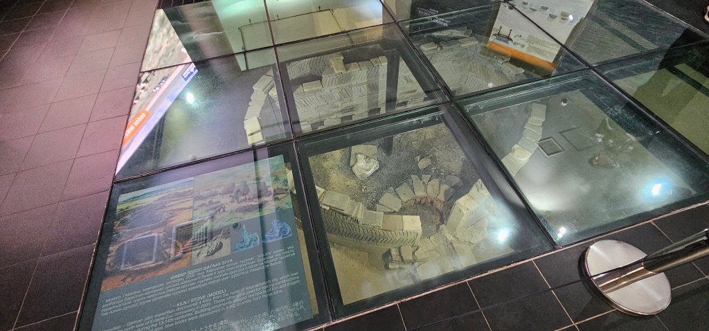 A glass display showing an archaeological site beneath, featuring stone structures and artifacts, with an informational panel nearby.
