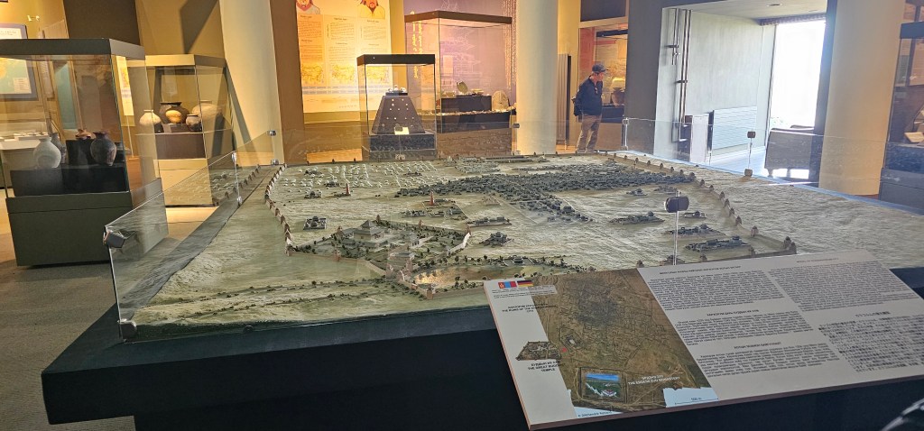 A detailed scale model of an archaeological site displayed in a museum, featuring buildings, roads, and landscape elements, with informational signage nearby.