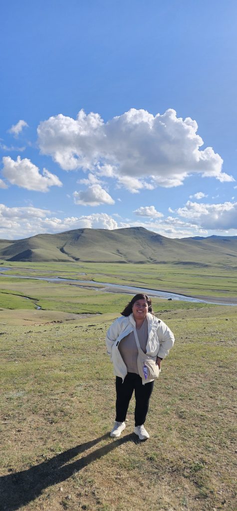 A person stands on a grassy hill with a picturesque landscape of rolling hills and a blue sky dotted with clouds in the background.