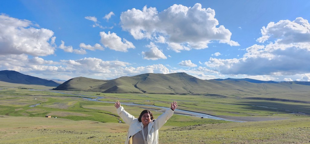 A person standing on a grassy hillside with arms raised, smiling against a backdrop of rolling green hills and a blue sky filled with fluffy clouds.