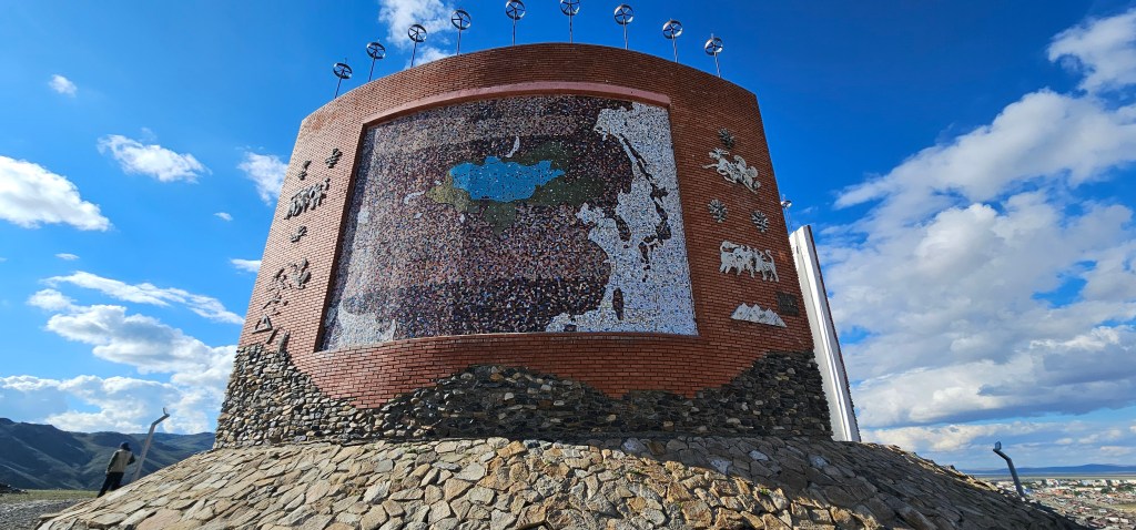 A large brick structure featuring a mosaic map of a region, with artistic elements such as horses and mountains, set against a clear blue sky with scattered clouds.