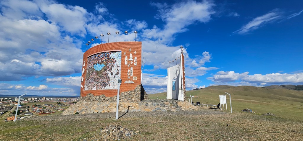 A large, colorful monument situated on a hill, featuring artwork and inscriptions, surrounded by a scenic landscape with blue skies and scattered clouds.
