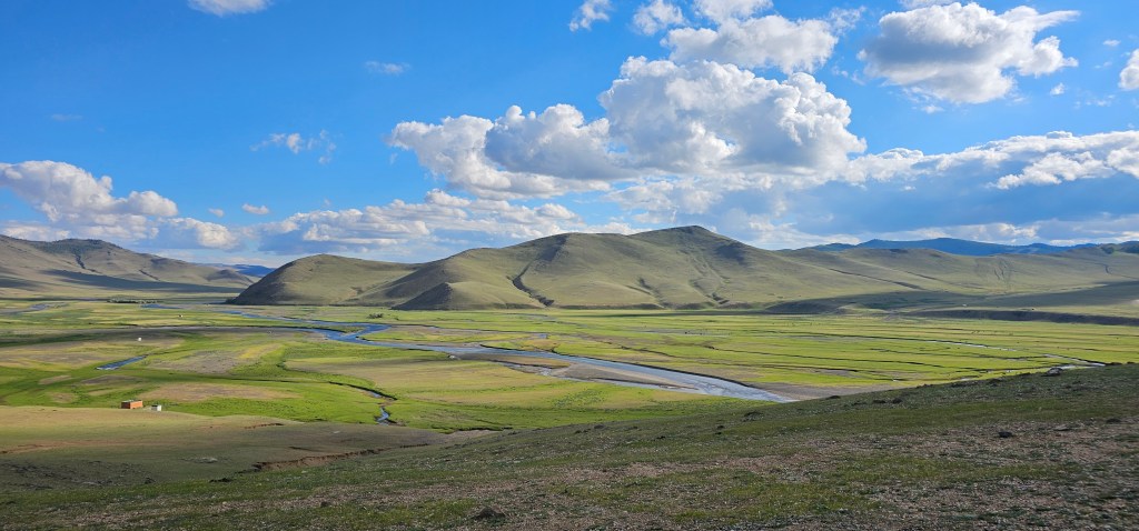 A panoramic view of a lush green landscape featuring rolling hills, a winding river, and a bright blue sky with fluffy white clouds.