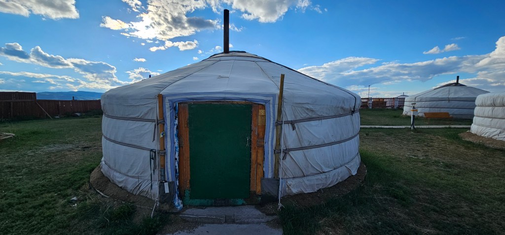 A close-up view of a traditional yurt with a green door, set against a backdrop of clouds and blue sky, with other yurts visible in the background.