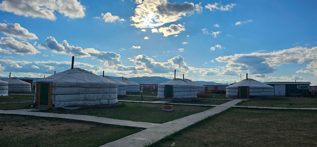 A scenic view of traditional yurts in a grassy area, under a blue sky filled with fluffy clouds, with mountains visible in the background.