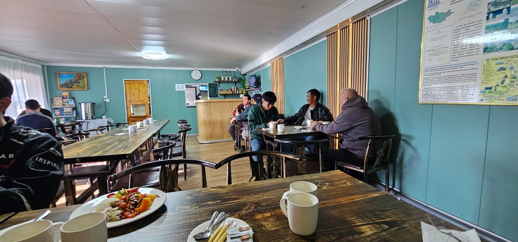 A cozy dining area with wooden tables and chairs, featuring a group of people dining and conversing, while a table in the foreground is set with plates of food and cups.